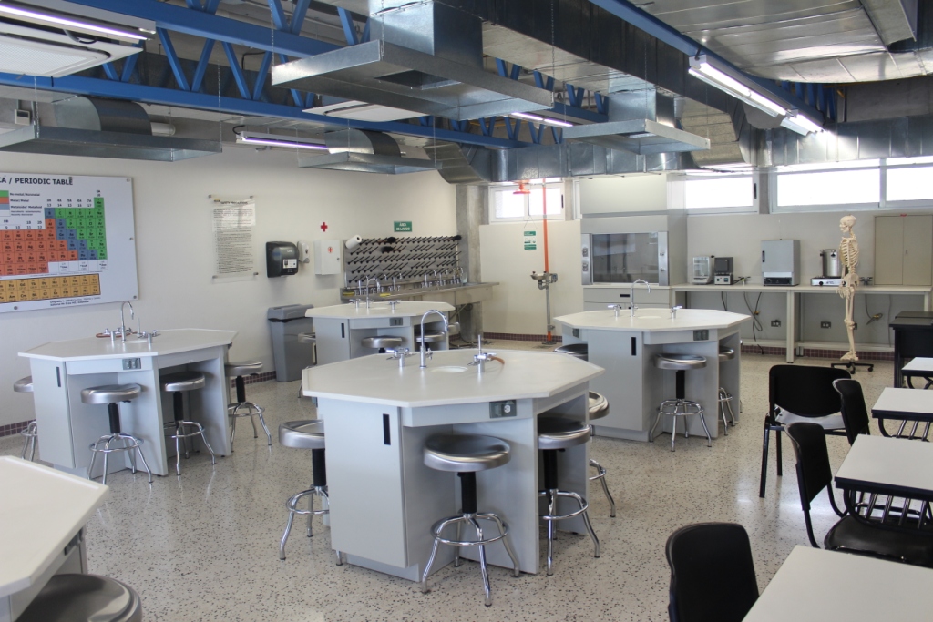 Laboratorio educativo equipado con mesas de trabajo hexagonales para estudiantes, tarjas integradas y campana de extracción al fondo.