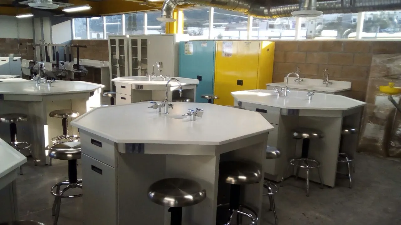 Laboratorio con mesas de trabajo, gabinetes de seguridad amarillo y azul, y estación de lavaojos